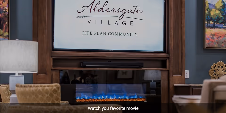 A cozy living room area featuring a modern electric fireplace with blue flames beneath a large screen displaying the text 'Aldersgate Village Life Plan Community.' The room includes a table lamp, upholstered chairs, and colorful artwork on the walls.
