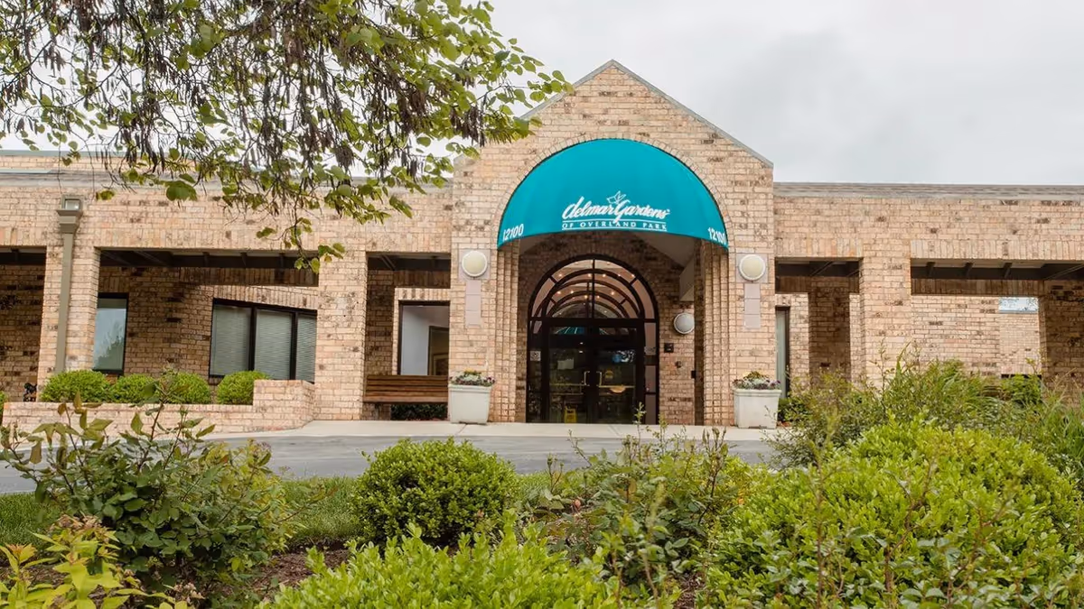 Front exterior view of Delmar Gardens of Overland Park building with a brick facade, green awning over the entrance, and surrounding greenery and bushes.