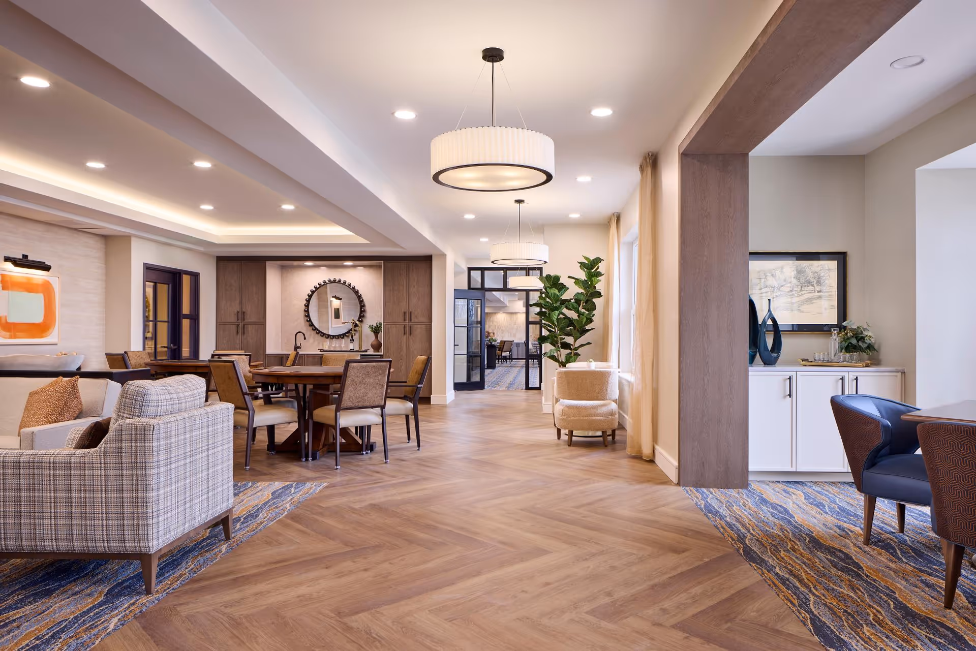 A spacious and well-lit common area in a senior living facility featuring a seating area with upholstered chairs and a sofa, a round wooden dining table with chairs, modern pendant lights, large windows with sheer curtains, decorative plants, and artwork on the walls. The flooring is a combination of wood and patterned carpet.