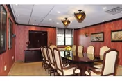 A formal dining room with a long wooden table surrounded by ten upholstered chairs. The walls are decorated with red patterned wallpaper and framed artwork. Two large windows allow natural light into the room, and two ornate ceiling light fixtures hang above the table.