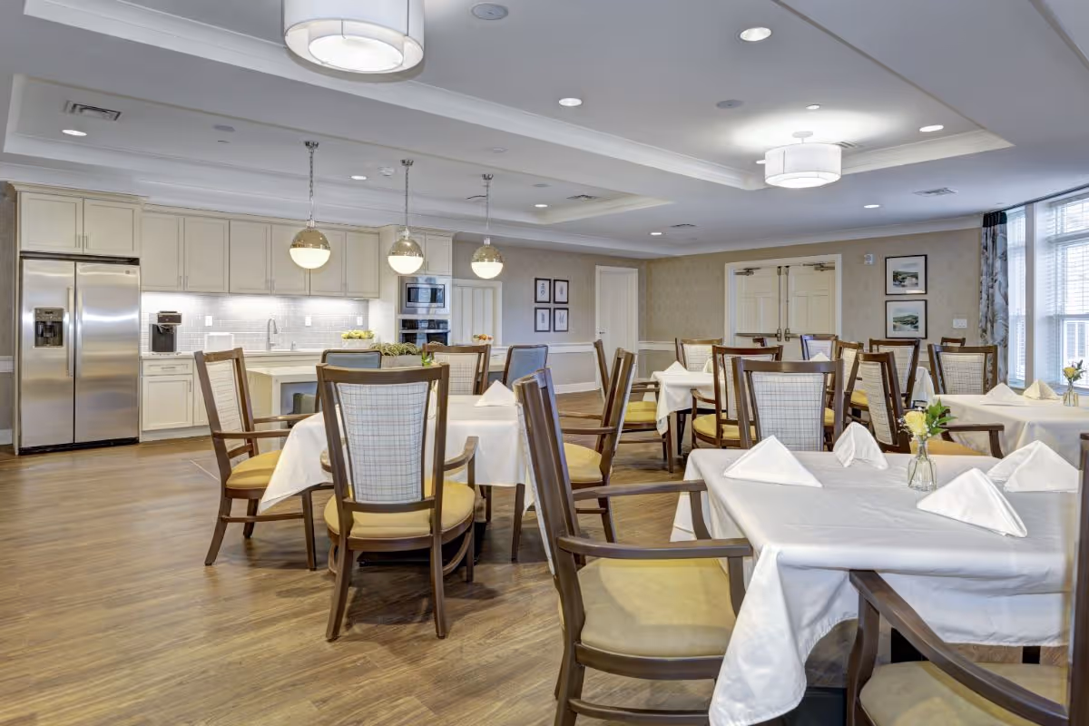 A bright dining room with multiple tables covered in white tablecloths and neatly folded napkins. Each table is surrounded by wooden chairs with cushioned seats. The room features a modern kitchen area with white cabinetry, stainless steel refrigerator, built-in oven, and pendant lights hanging over the counter. The walls are decorated with framed pictures, and large windows allow natural light to fill the space.