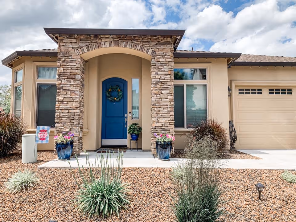 Front exterior view of a single-story house with a stone archway entrance, a blue front door decorated with a wreath, two large windows, a beige garage door, and a landscaped front yard with gravel and various plants.