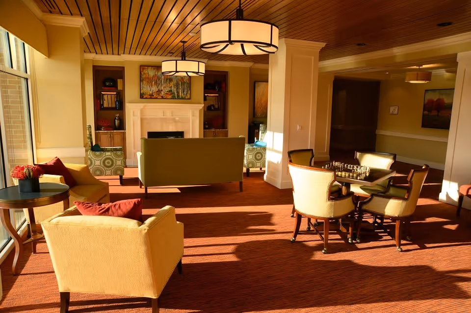 A cozy senior living facility common area with comfortable armchairs and sofas arranged around a fireplace. There is a round table with four chairs and a chessboard set up. The room has large windows letting in natural light, warm-colored carpet, wooden ceiling, and modern hanging light fixtures. Artwork and shelves decorate the walls near the fireplace.