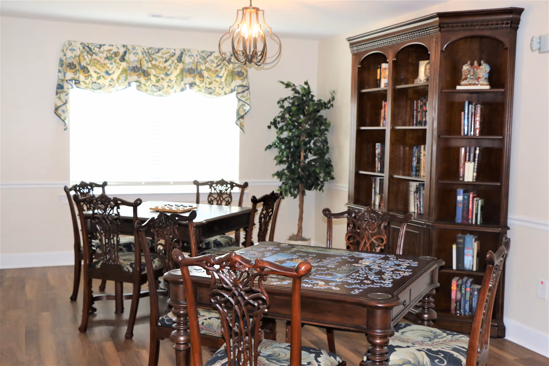 A bright room with two wooden tables, each surrounded by four ornate wooden chairs with patterned cushions. One table has a partially completed jigsaw puzzle on it. There is a large wooden bookshelf filled with books and decorative items against the wall. A large window with floral valance curtains lets in natural light, and a potted artificial tree stands in the corner. The floor is wooden, and a decorative ceiling light fixture hangs above.