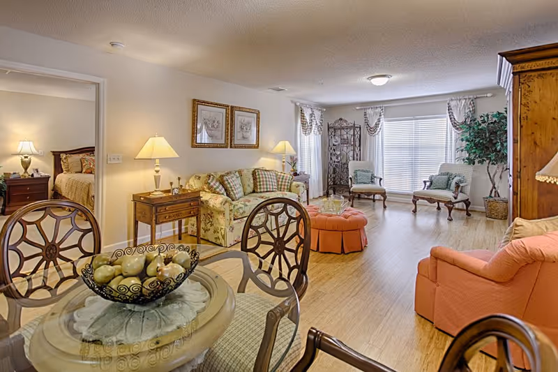 A bright and spacious senior living facility interior featuring a living room with floral and plaid upholstered sofa, two armchairs, a coral-colored armchair, and a round glass-top dining table with decorative chairs. The room has wooden flooring, two table lamps, framed artwork on the wall, and large windows with sheer curtains allowing natural light. A bedroom with a bed and nightstand is visible through an open doorway.