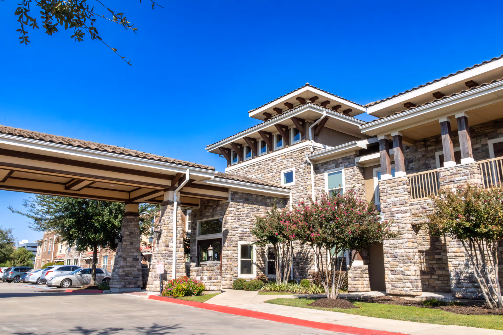 Exterior view of a senior living facility building with stone facade, a covered entrance, landscaped bushes, and parked cars under a clear blue sky.