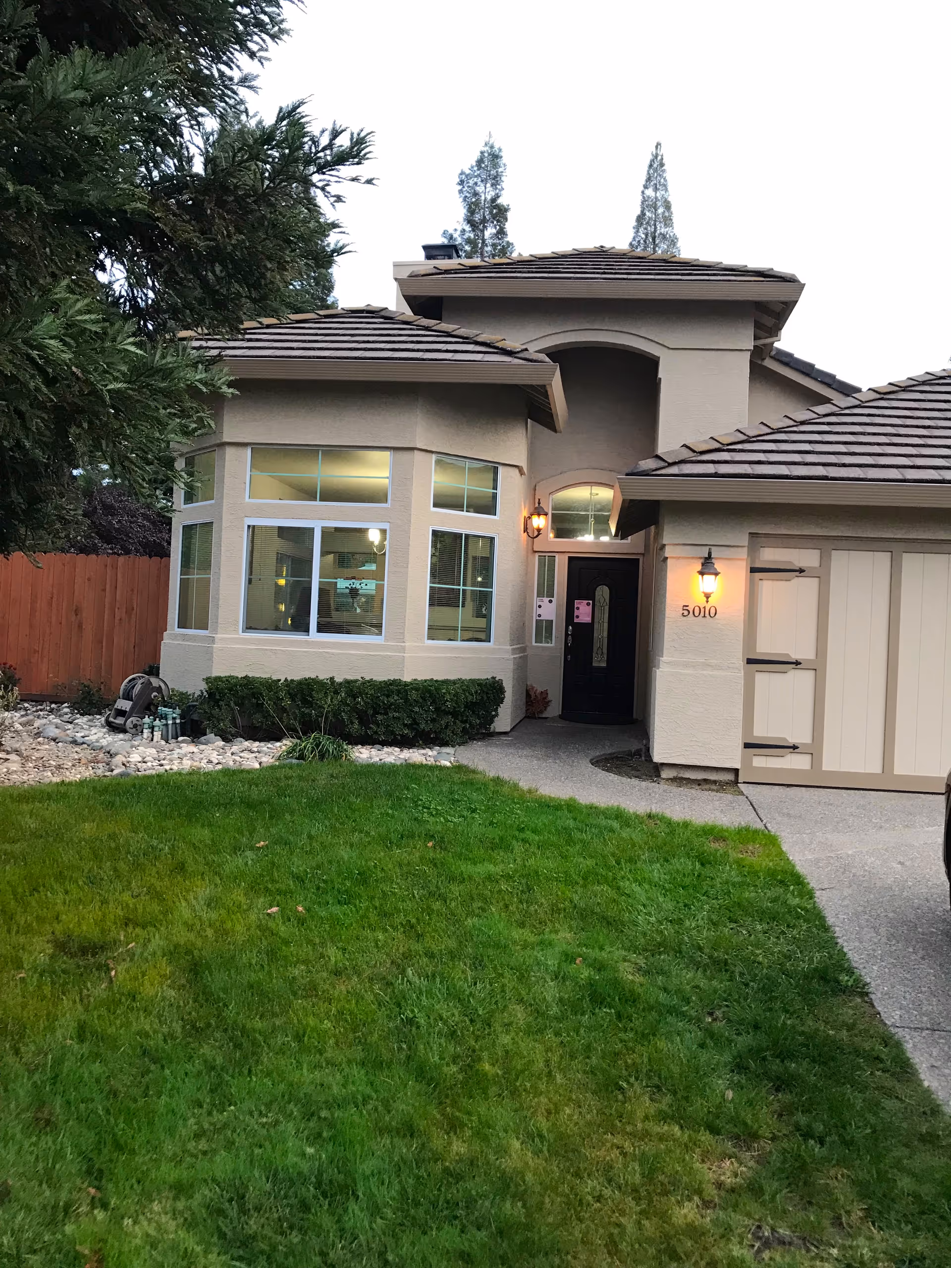 Front exterior view of a single-story beige house with a well-maintained green lawn, a pathway leading to a black front door with a light fixture above it, and a garage door on the right side. There are large windows on the left side of the entrance and some bushes along the house. Trees and a wooden fence are visible in the background.
