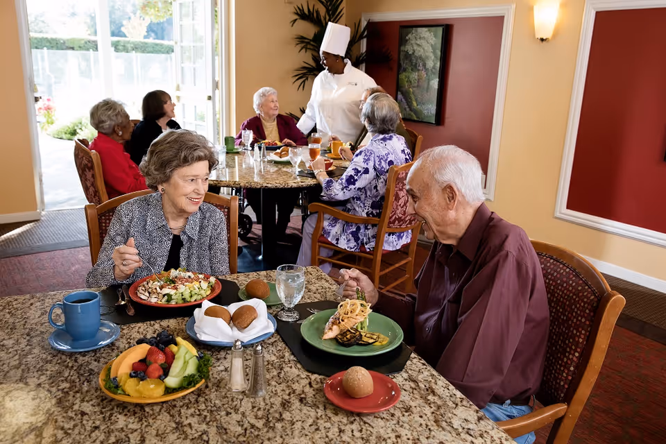 Several elderly people enjoying a meal together in a dining room at a senior living facility. A chef in a white uniform is standing and interacting with the group. The table is set with plates of food, drinks, and bread rolls. The room has warm-colored walls and large windows letting in natural light.