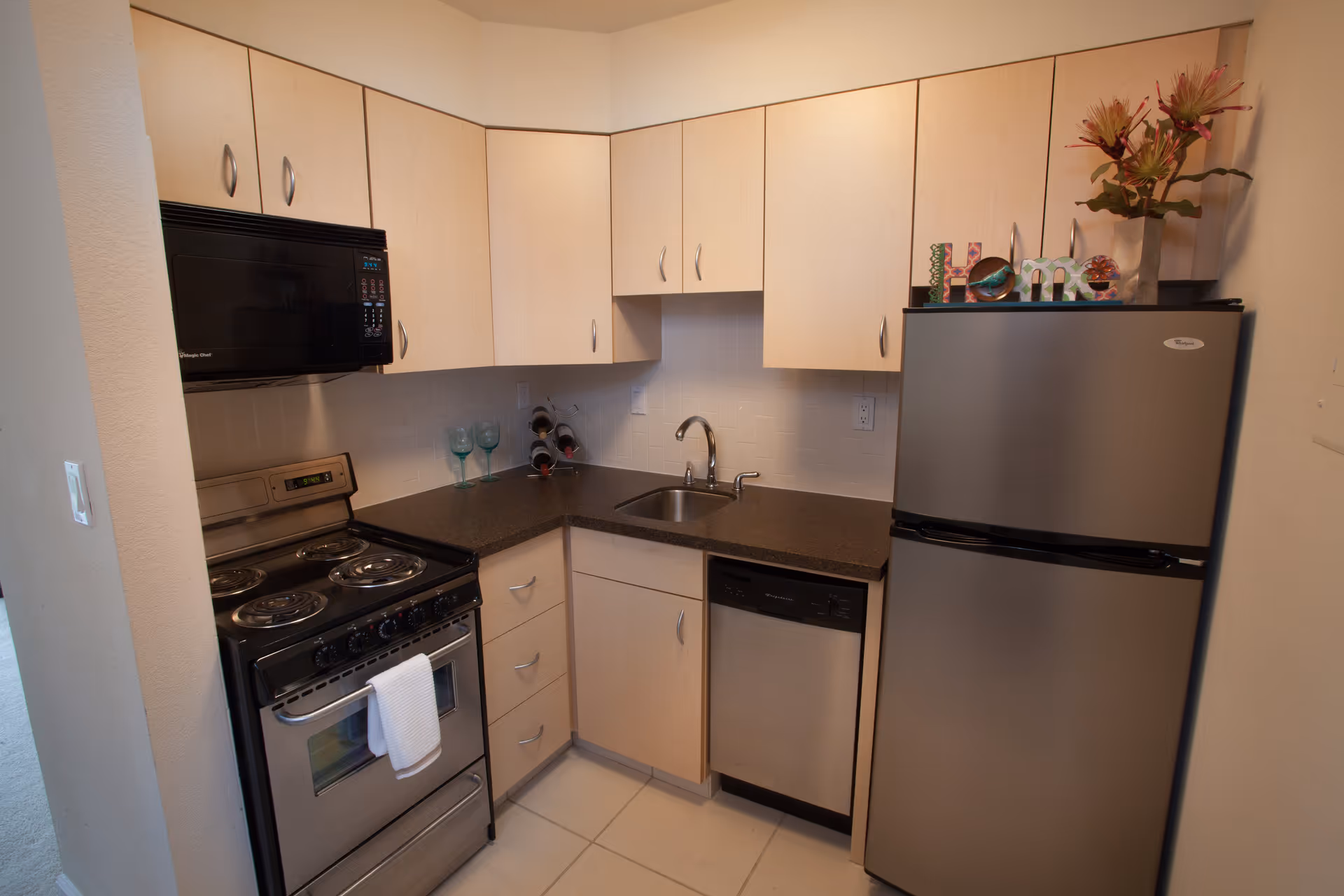 Compact kitchen with light wood cabinets, black countertop, stainless steel refrigerator, dishwasher, electric stove with oven, and a black microwave. There are three wine glasses and a small wine rack on the counter, and a decorative 'Home' sign with flowers on top of the refrigerator.
