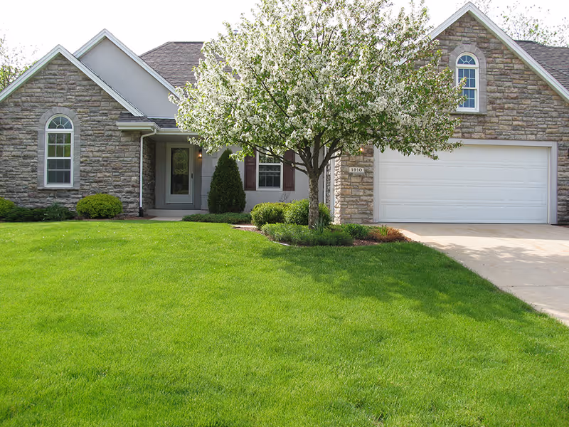 Front exterior view of a single-story house with stone and siding facade, a white garage door, a blossoming tree, and a well-maintained green lawn.