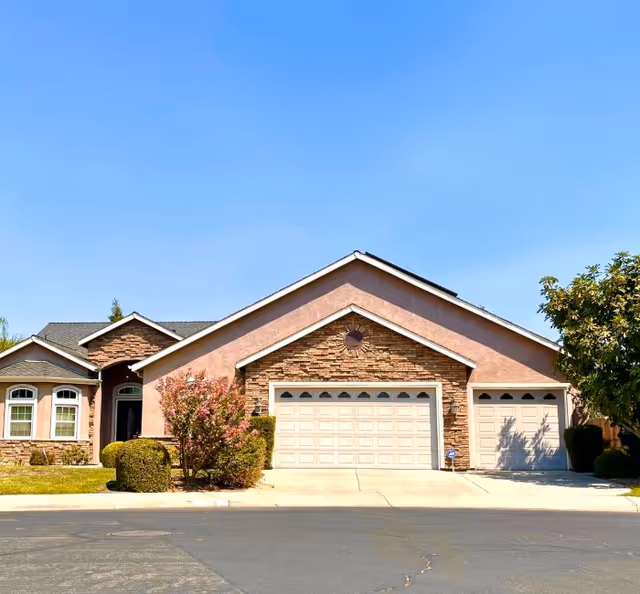 Front exterior view of a single-story house with a three-car garage, stone and stucco facade, manicured bushes, and a clear blue sky.