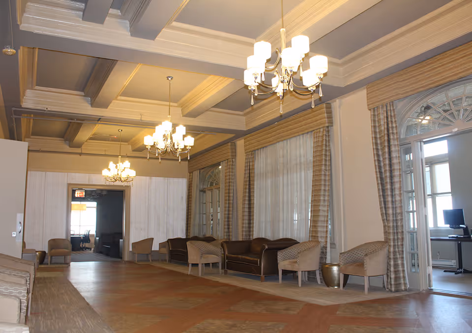 A spacious, well-lit common area with multiple chandeliers hanging from a coffered ceiling. The room features large windows with sheer curtains and striped drapes, several armchairs, and a brown leather sofa arranged along the walls. There is an open doorway leading to another room with office furniture visible.
