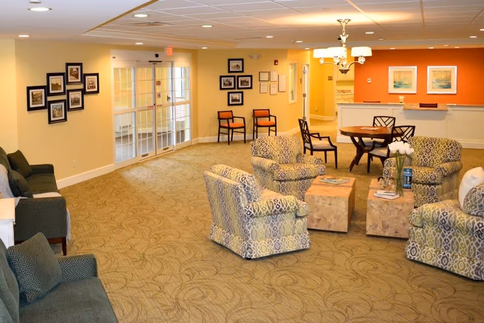 A spacious and well-lit common area in a senior living facility with patterned armchairs arranged around two wooden coffee tables, a round table with chairs, and a reception desk with orange accent wall behind it. The room has beige walls, framed pictures on the walls, and a carpeted floor with a subtle pattern.