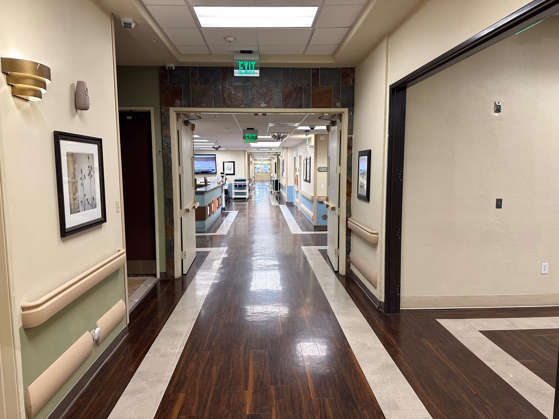 A clean, well-lit hallway in a nursing and rehabilitation center with polished wooden floors bordered by light-colored tiles. The hallway has handrails on both sides, framed artwork on the walls, and double doors leading to another corridor with medical carts and monitors visible in the distance. Exit signs are mounted on the ceiling.