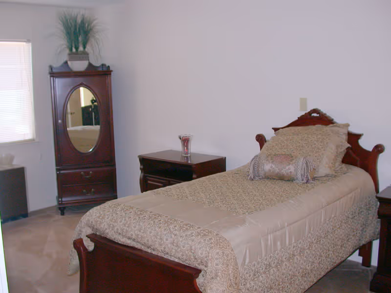 A bedroom with a single wooden bed covered with a beige patterned bedspread and matching pillows. Next to the bed is a wooden nightstand with a glass vase on top. In the corner of the room is a tall wooden cabinet with an oval mirror and a potted plant on top. A window with closed blinds is visible on the left side.