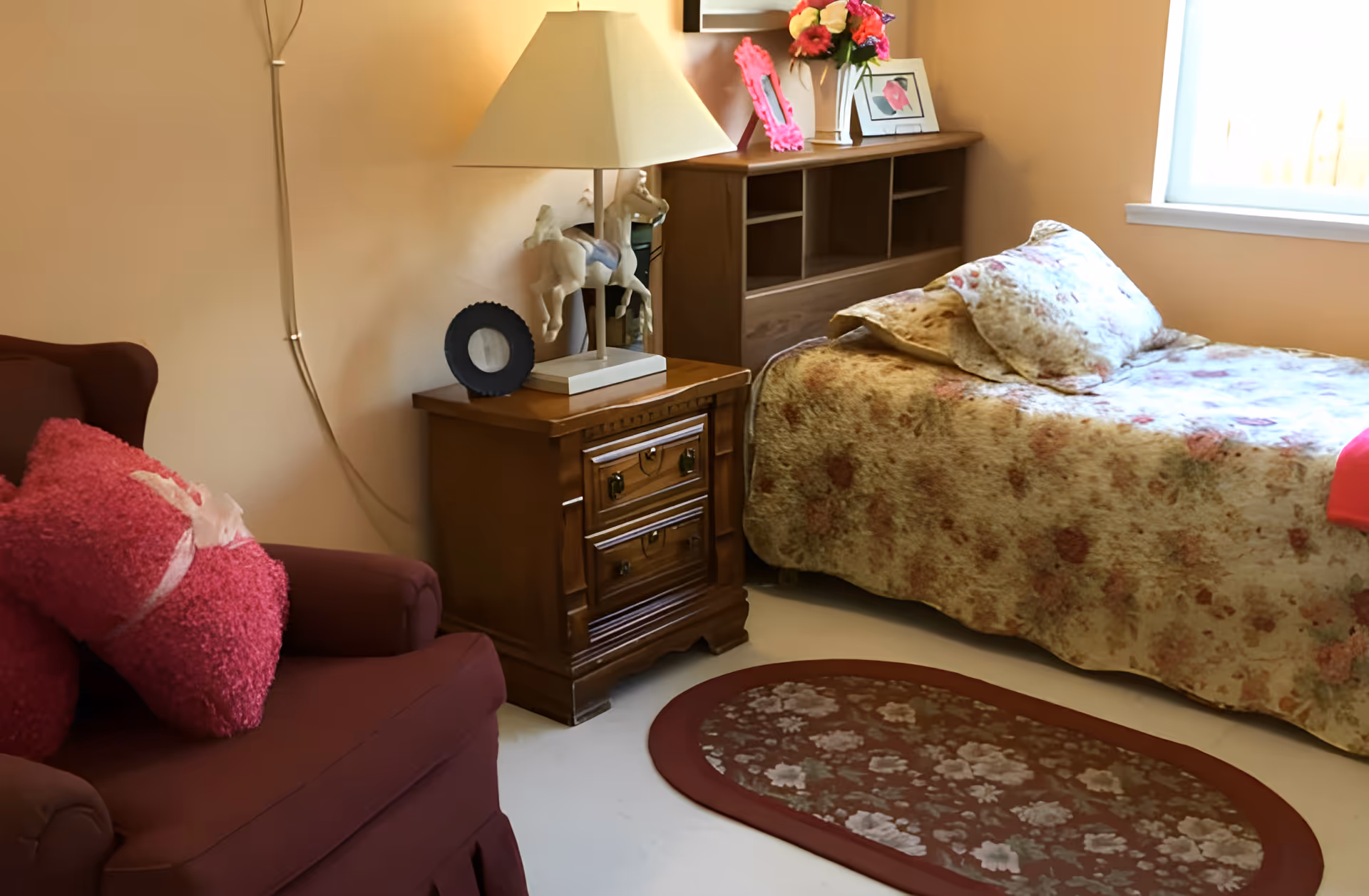 A cozy bedroom with a single bed covered in a floral-patterned blanket and matching pillow. Next to the bed is a wooden nightstand with two drawers, a lamp with a horse-shaped base, and decorative items including a photo frame and a vase with flowers. A maroon armchair with pink cushions is positioned to the left, and a floral oval rug lies on the floor. A window on the right side lets in natural light.
