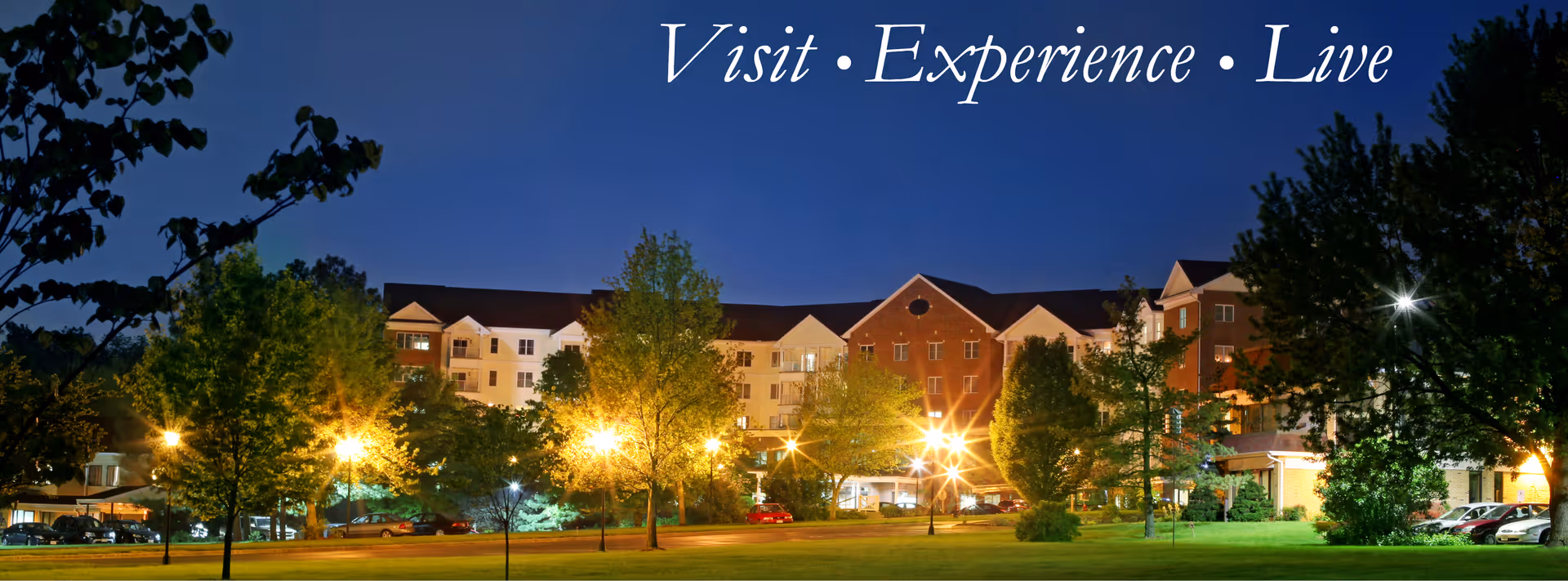 Nighttime view of a large, well-lit retirement community building surrounded by trees and a grassy area, with the words 'Visit • Experience • Live' displayed at the top.