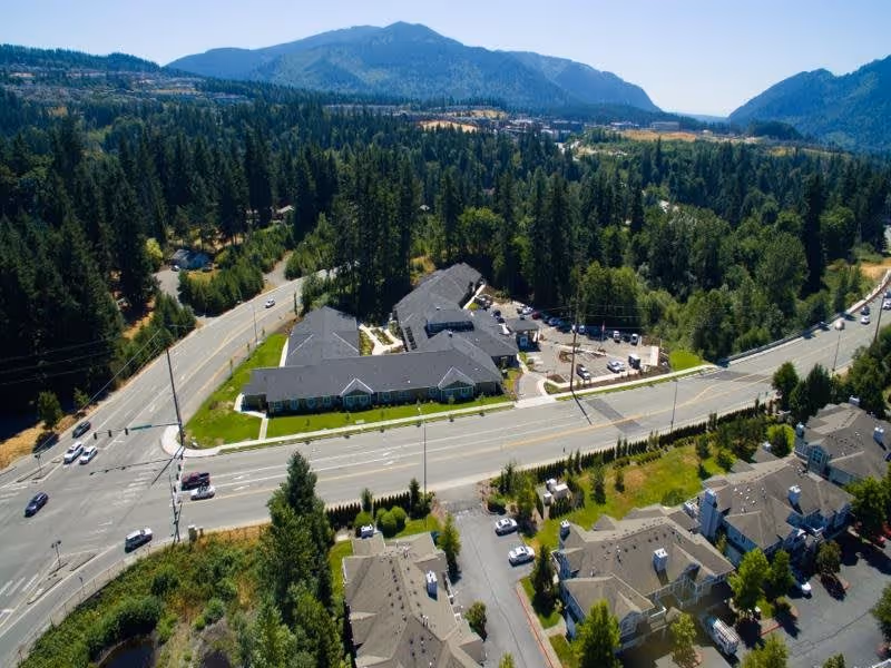 Aerial view of a senior living facility named Ciel of Issaquah surrounded by trees and mountains in the background. The facility buildings are situated near a main road with cars and parking areas visible. Residential houses are also seen nearby.