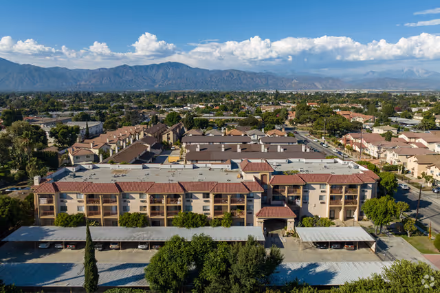 Aerial view of Flamingo Garden Apartments, showing multiple residential buildings with red-tiled roofs, covered parking areas, and surrounding trees. In the background, there is a sprawling suburban neighborhood with mountains under a partly cloudy sky.