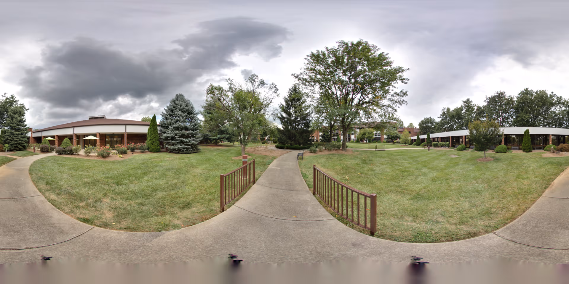 Outdoor view of a senior living facility with a concrete pathway leading through a grassy area with trees and shrubs. There are buildings on both sides with large windows and a cloudy sky overhead.