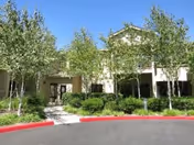 Exterior view of a senior living facility building partially obscured by several leafy green trees and shrubs, with a paved driveway and red-painted curb in the foreground.