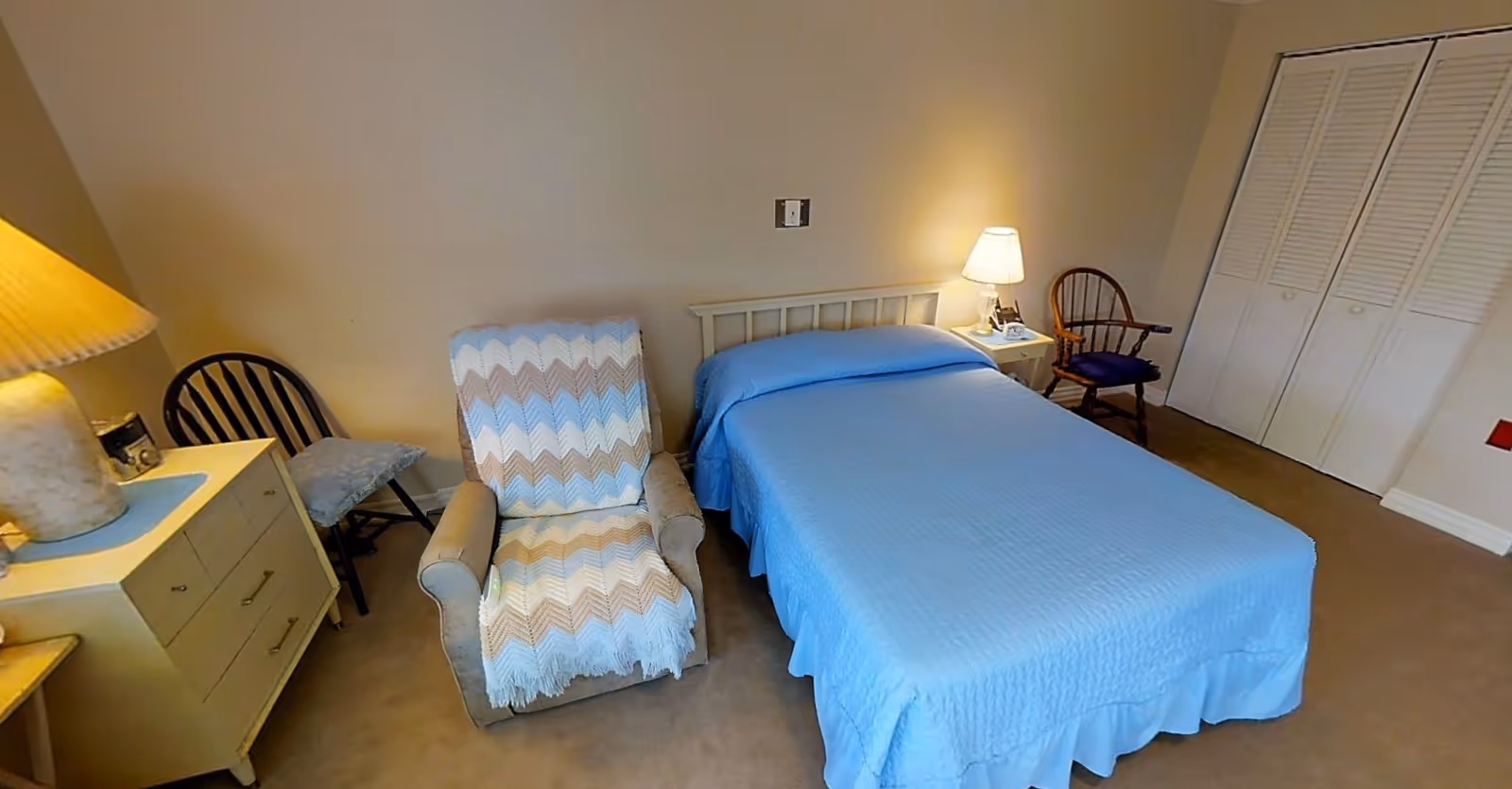 A bedroom with a bed covered in a blue quilt, a beige armchair with a chevron-patterned throw, two wooden chairs, a nightstand with a lamp, and a dresser with a lamp on it. There is a closet with louvered doors on the right side of the room.