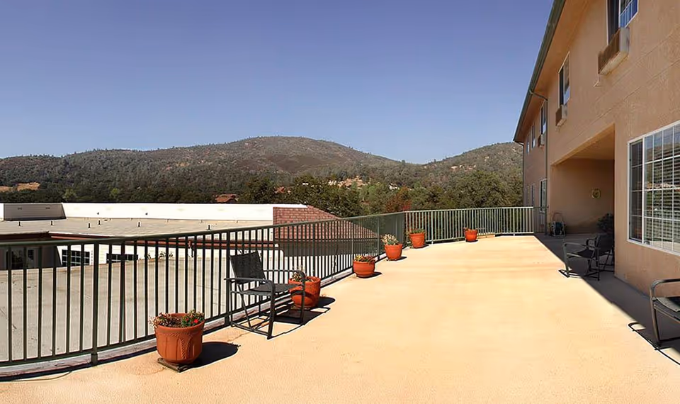 Outdoor patio area at Skyline Place Senior Living with several potted plants along a metal railing, a few chairs, and a view of distant hills under a clear blue sky.