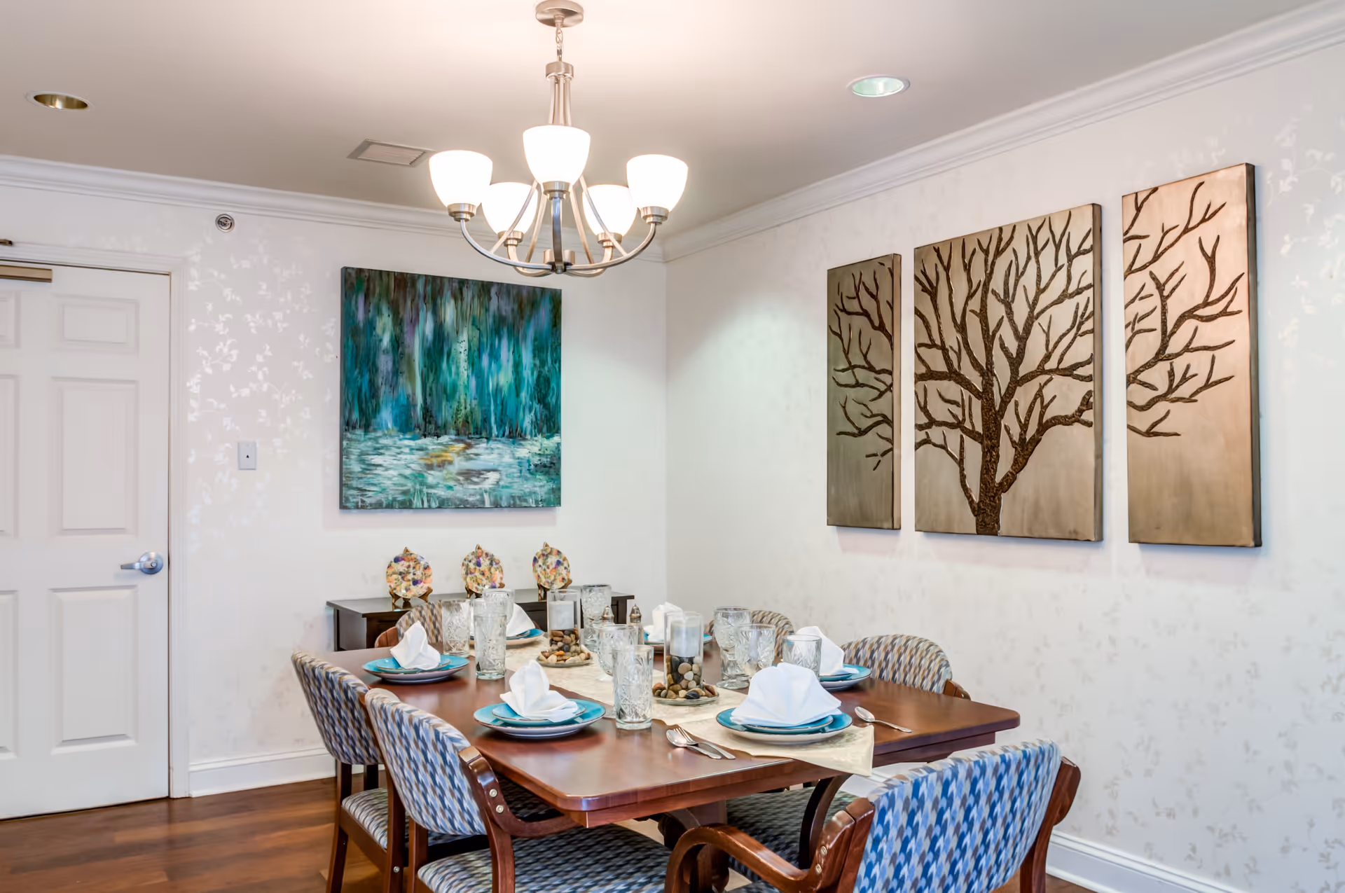 A dining room with a wooden table set for six people with plates, napkins, glasses, and silverware. The chairs have patterned upholstery. On the wall, there are three-panel artwork of a tree and another painting with abstract green and blue colors. A chandelier with five lights hangs above the table.
