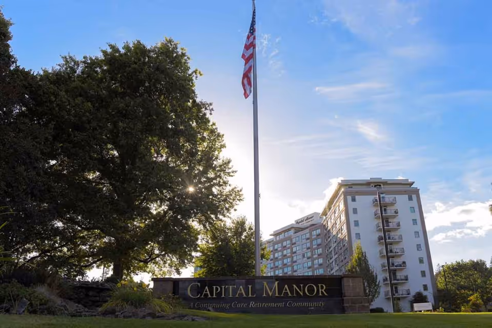 Exterior view of Capital Manor, a continuing care retirement community, showing a multi-story building with balconies, a large tree, an American flag on a flagpole, and a sign with the community name in front.