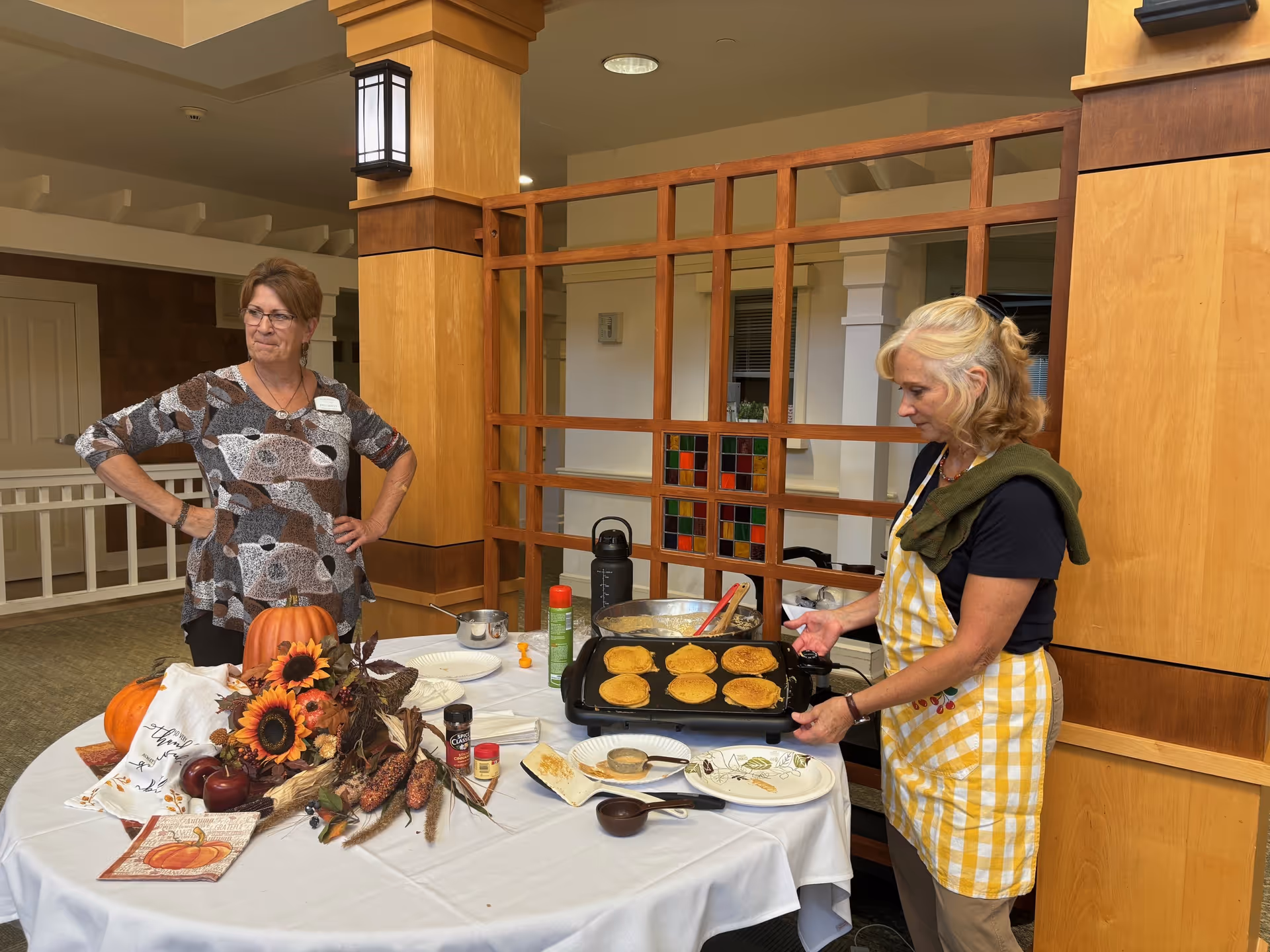 Two women in an indoor setting at Maplewood at Chardon. One woman wearing a patterned top stands with her hands on her hips near a table decorated with autumn-themed items including pumpkins, sunflowers, and corn. The other woman, wearing a yellow and white checkered apron, is cooking pancakes on an electric griddle placed on the table. The background features wooden pillars and a wooden lattice partition.