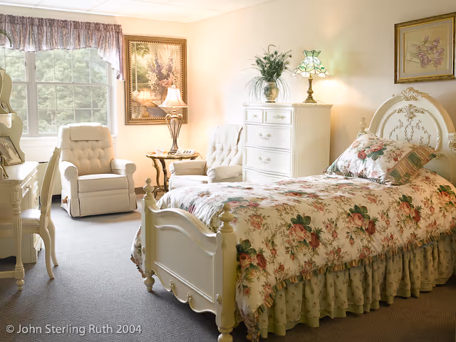 A cozy bedroom with a floral-patterned bedspread on a white ornate bed frame. The room features two cushioned armchairs near a window with floral curtains, a white dresser with a decorative lamp and plant on top, and framed artwork on the walls. The room is softly lit with natural and lamp light.