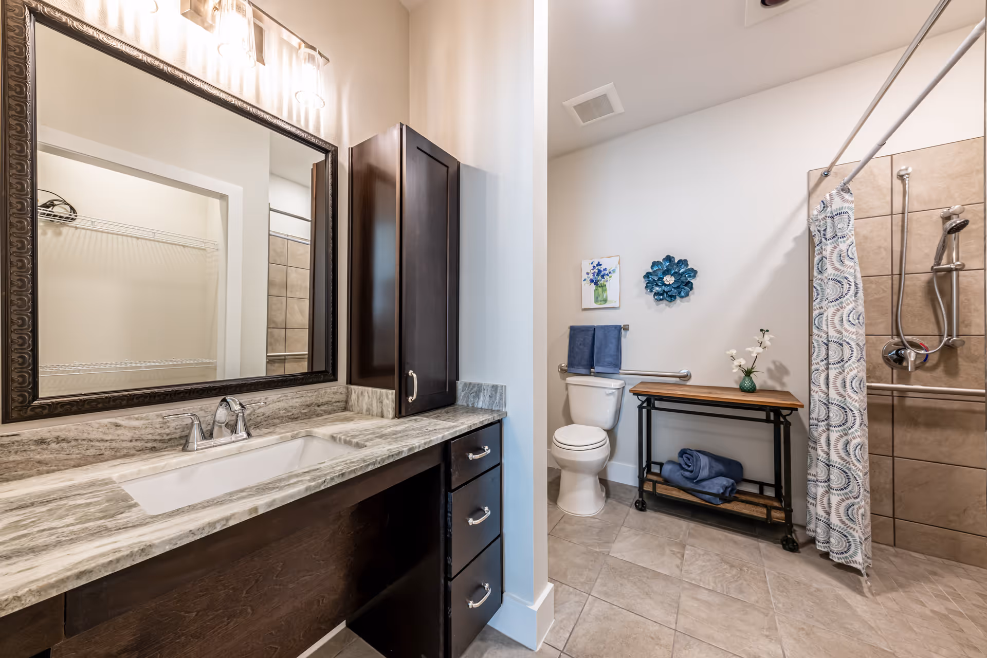 A modern bathroom featuring a large mirror above a marble countertop with a sink and dark wood cabinetry. The room includes a toilet with a towel rack above it, a decorative blue flower wall art, a small table with a vase of flowers and folded towels, and a walk-in shower with a patterned shower curtain and grab bars.