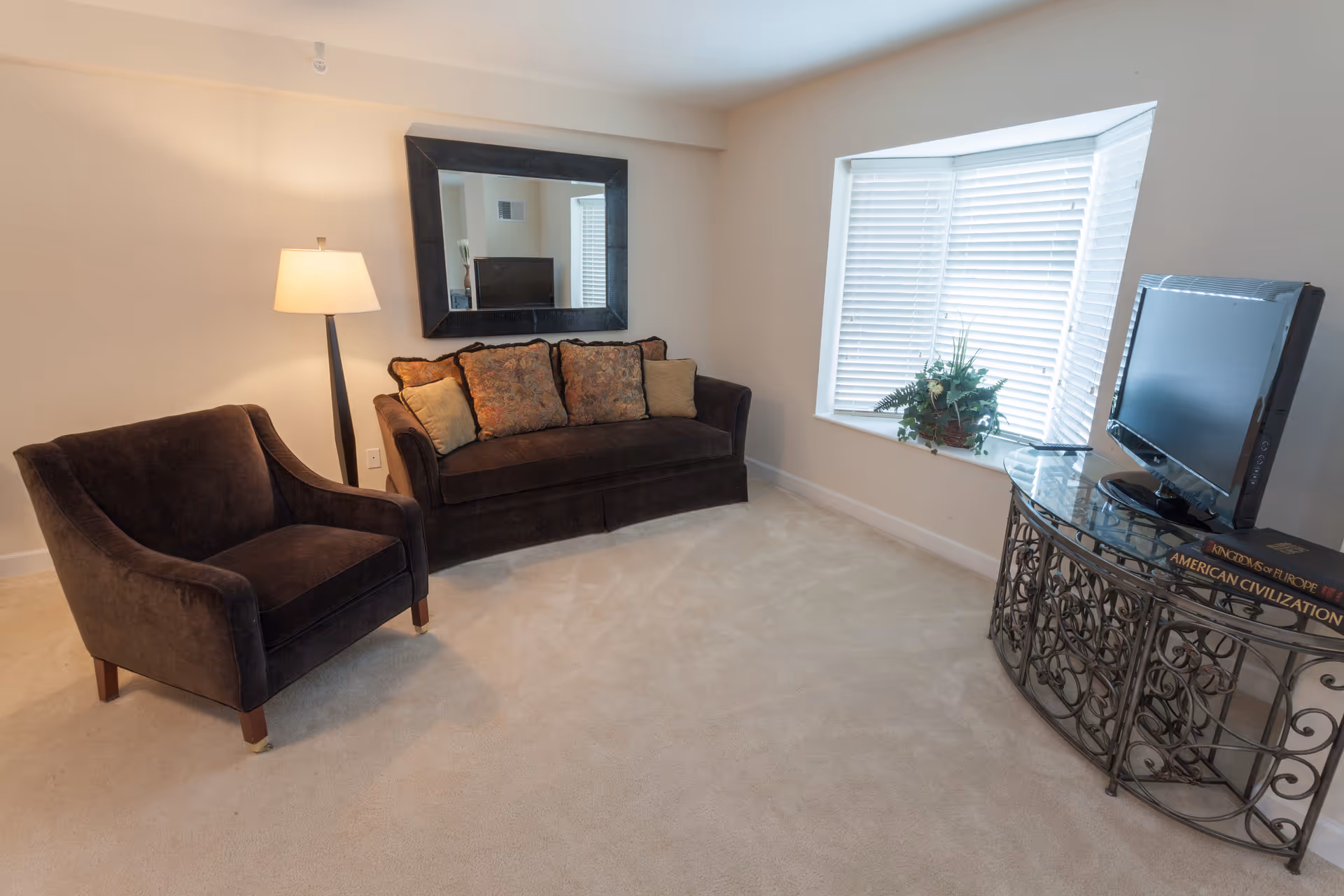 A cozy living room with a dark brown armchair and matching sofa adorned with patterned cushions. A floor lamp stands beside the sofa, and a large mirror hangs on the wall above it. A bay window with white blinds lets in natural light, and a decorative plant sits on the windowsill. A flat-screen TV is placed on an ornate metal and glass table, which also holds two large books.