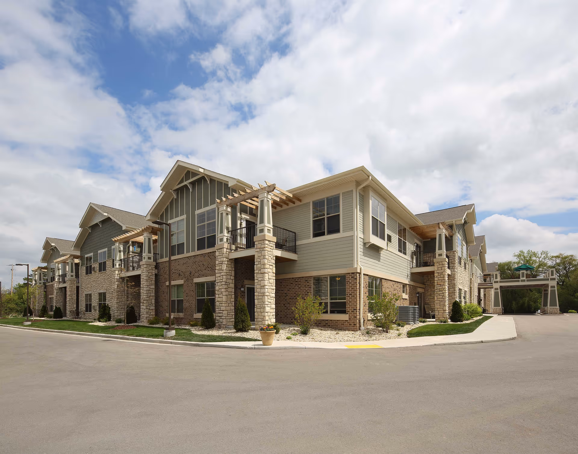Exterior view of a two-story assisted living facility building with a combination of brick and siding walls, multiple windows, balconies with pergolas, and a paved driveway under a partly cloudy sky.