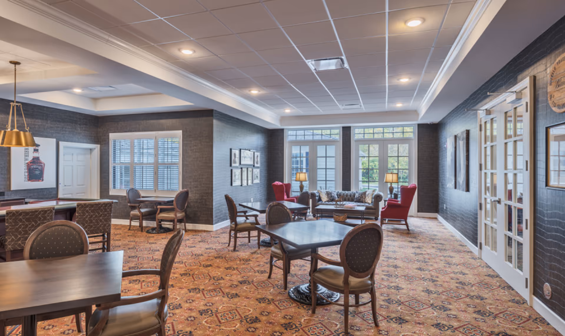 A spacious senior living common area with multiple tables and chairs arranged for socializing or activities. The room features a patterned carpet, dark textured walls, and a ceiling with recessed lighting. In the background, there is a seating area with a sofa and two red armchairs near large windows and glass doors letting in natural light. French doors are visible on the right side of the room.