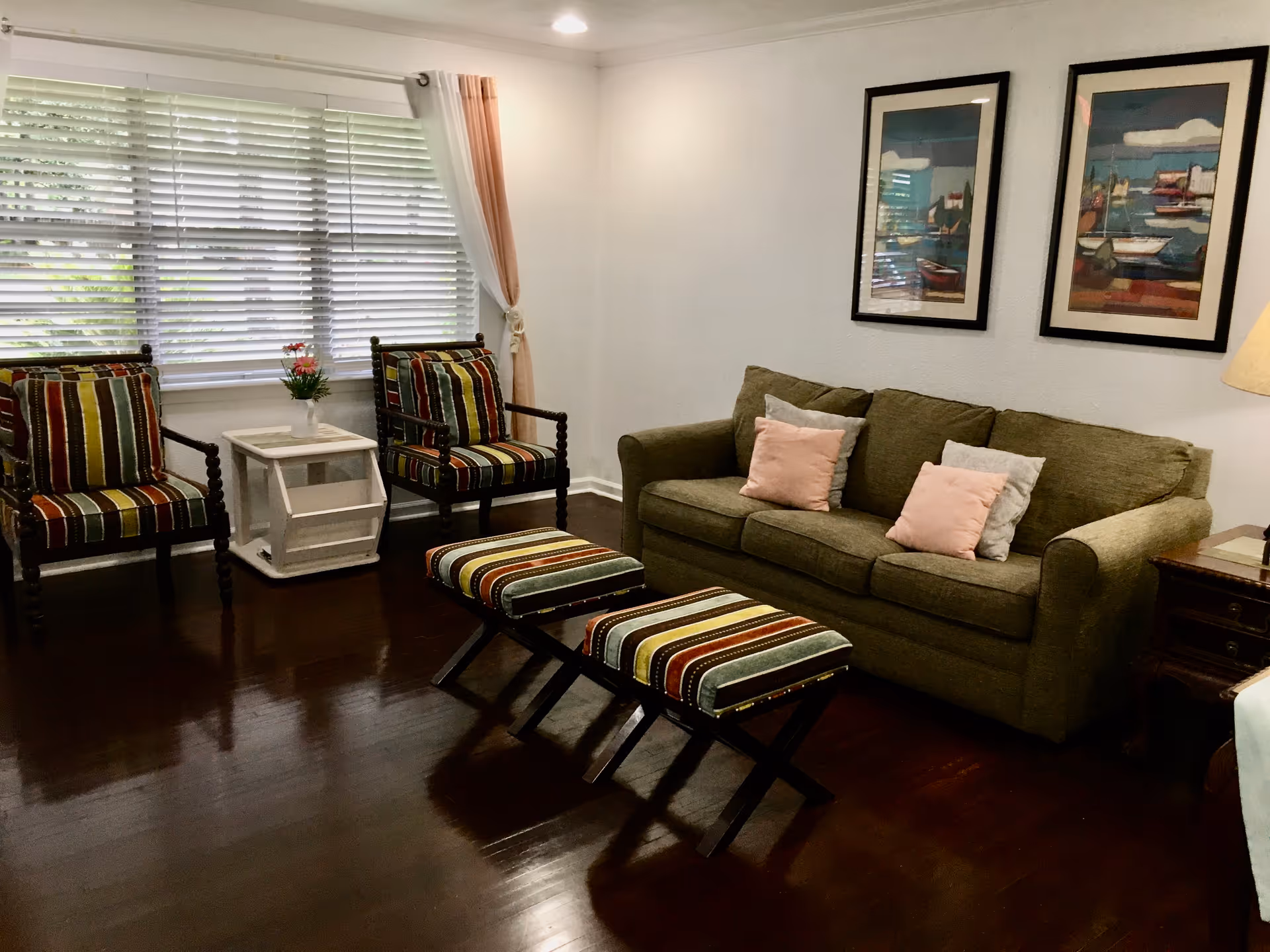A cozy living room with a green sofa adorned with pink and gray pillows, two striped armchairs, and two matching striped ottomans. A small white side table with a vase of flowers is placed between the armchairs. The room has dark wooden floors, a large window with white blinds and peach-colored curtains, and two framed paintings of boats on the wall above the sofa.