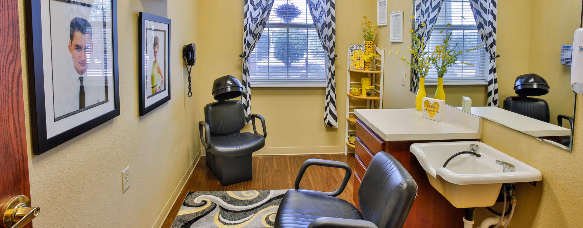 A small salon room with two black salon chairs, a white sink attached to a wooden cabinet, a large mirror, two windows with black and white chevron curtains, framed pictures on the wall, and a small shelf with yellow decorative items.