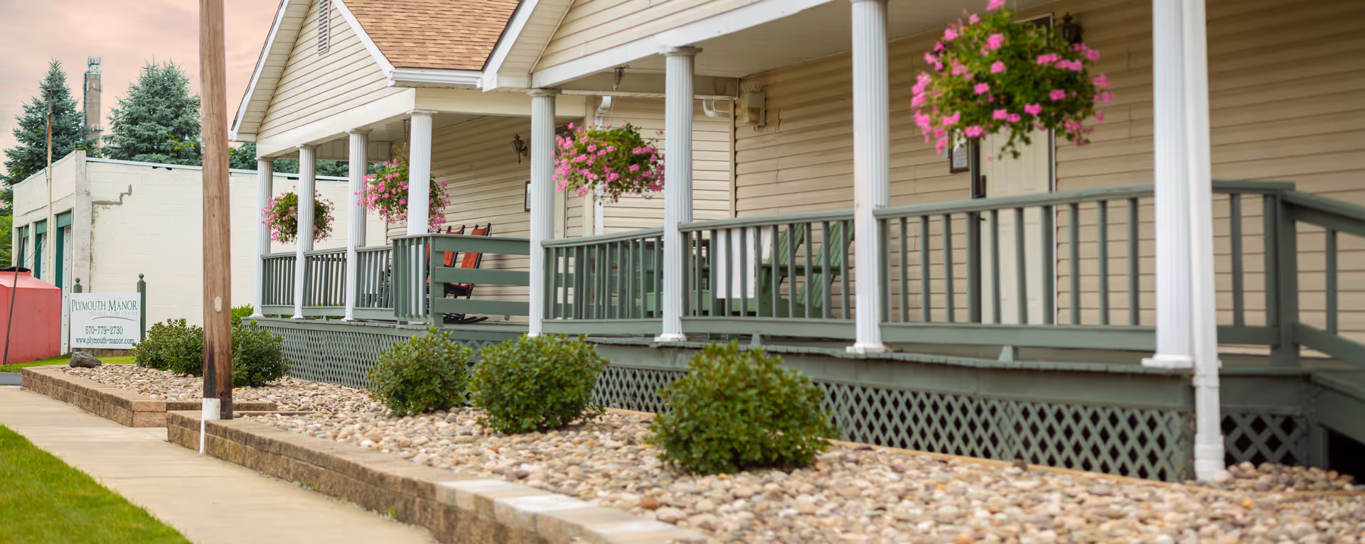 Exterior view of Plymouth Manor Personal Care Center showing a beige building with a green porch railing, white columns, hanging flower baskets, and a landscaped area with bushes and rocks along the sidewalk.