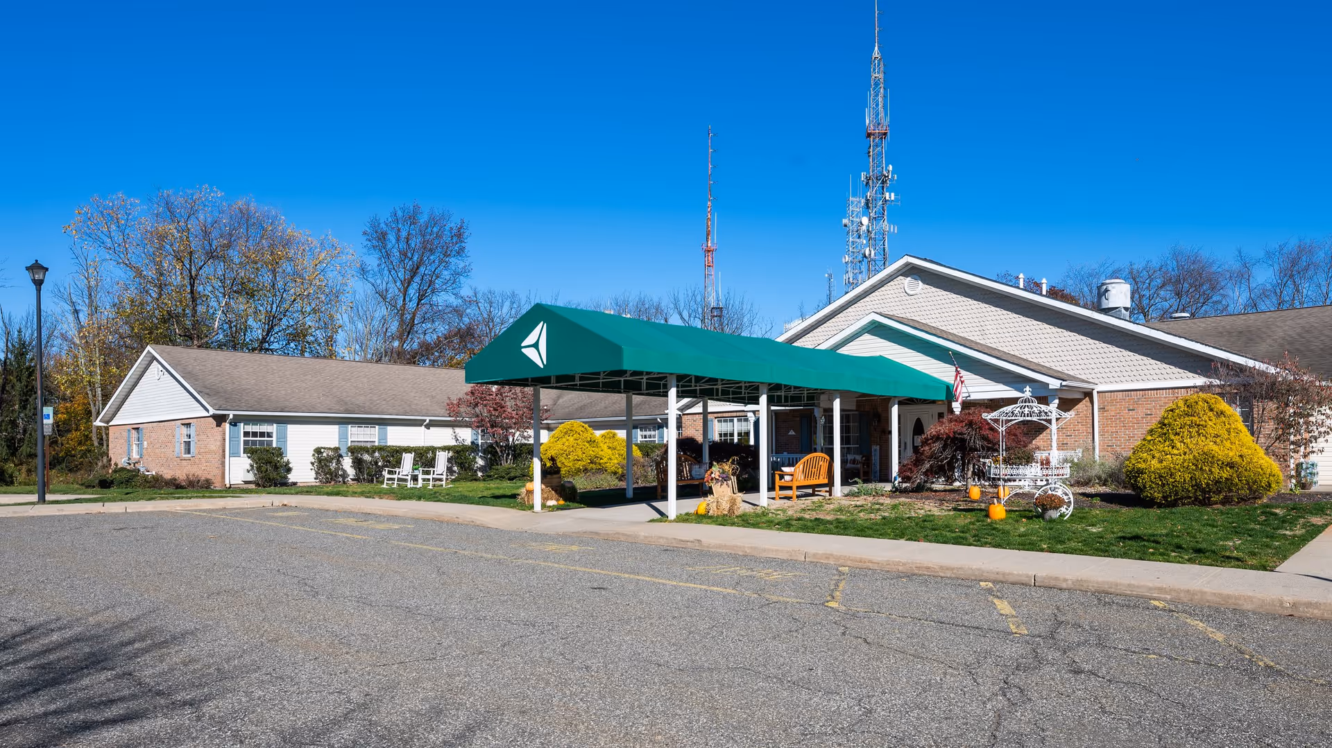 Exterior view of Arden Courts – ProMedica Memory Care Community (West Orange) showing a single-story building with a green canopy over the entrance, a paved parking area in front, landscaped bushes, and clear blue sky.