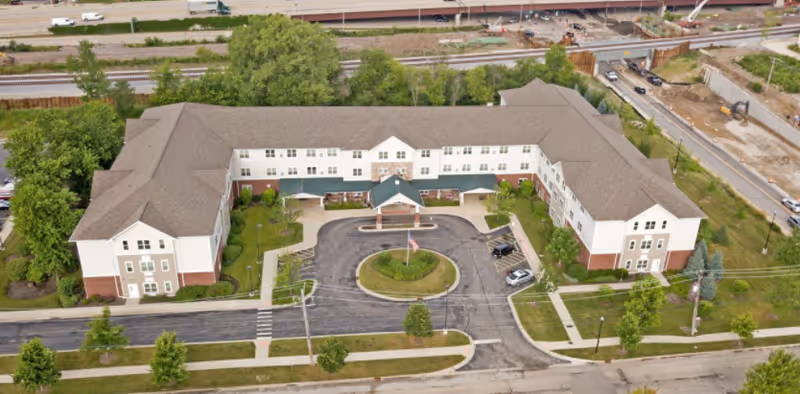 Aerial view of a large senior living facility building with a U-shaped layout, surrounded by greenery and roads. The building has a covered entrance with a circular driveway and an American flag in the center. Nearby are train tracks and a highway overpass.