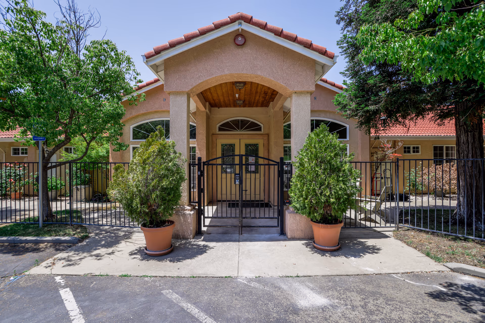 Front entrance of a senior living facility with a covered porch, double doors, and a black metal gate. Two large potted plants flank the entrance, and there are trees and greenery on either side of the building. The building has a beige exterior with a red-tiled roof.