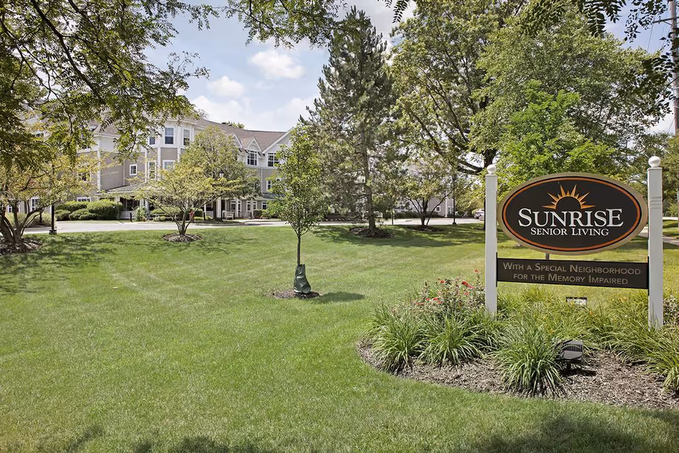 Exterior view of Sunrise Senior Living facility with a large green lawn, trees, and a sign that reads 'Sunrise Senior Living - With a Special Neighborhood for the Memory Impaired'. The building is visible in the background under a partly cloudy sky.