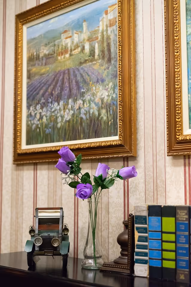 A decorative interior scene featuring a small vintage toy car, a clear glass vase with purple artificial flowers, a row of books with colorful spines, and a framed painting of a countryside landscape with fields and buildings on a wallpapered wall with vertical stripes.