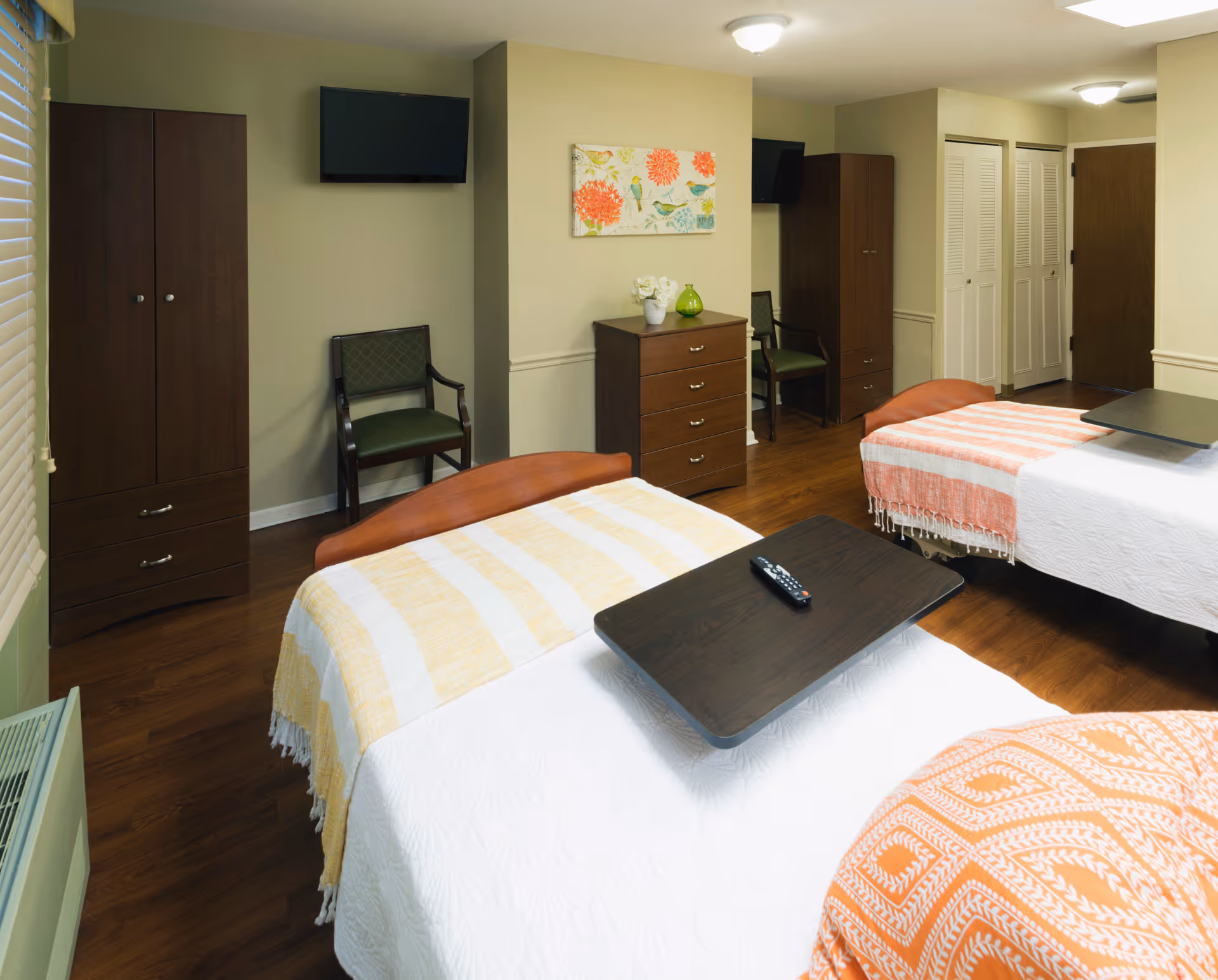 A senior living facility bedroom with two single beds, each covered with white bedspreads and colorful throws. Each bed has a dark wooden overbed table, one with a remote control on it. The room features wooden flooring, two dark wooden wardrobes, a chest of drawers with decorative items on top, two chairs, a wall-mounted TV, and a colorful floral painting on the wall.
