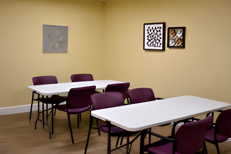 A room with two white rectangular tables and several purple chairs arranged around them. The walls are painted beige and have three abstract paintings hanging on them.