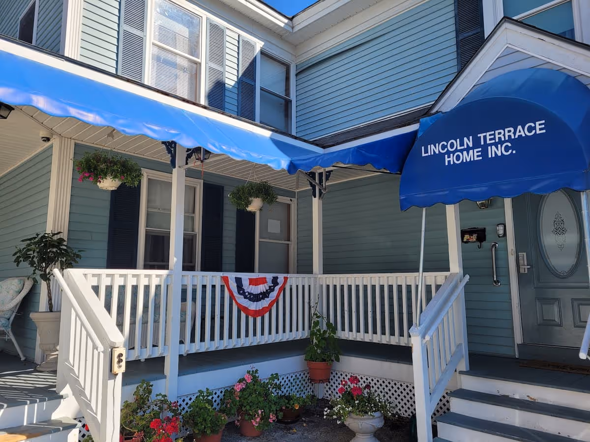Exterior view of a light blue building with white railings and stairs leading to a door. A blue awning above the door reads 'LINCOLN TERRACE HOME INC.'. There are hanging flower pots and several potted plants along the porch area, with a red, white, and blue decorative banner on the railing.