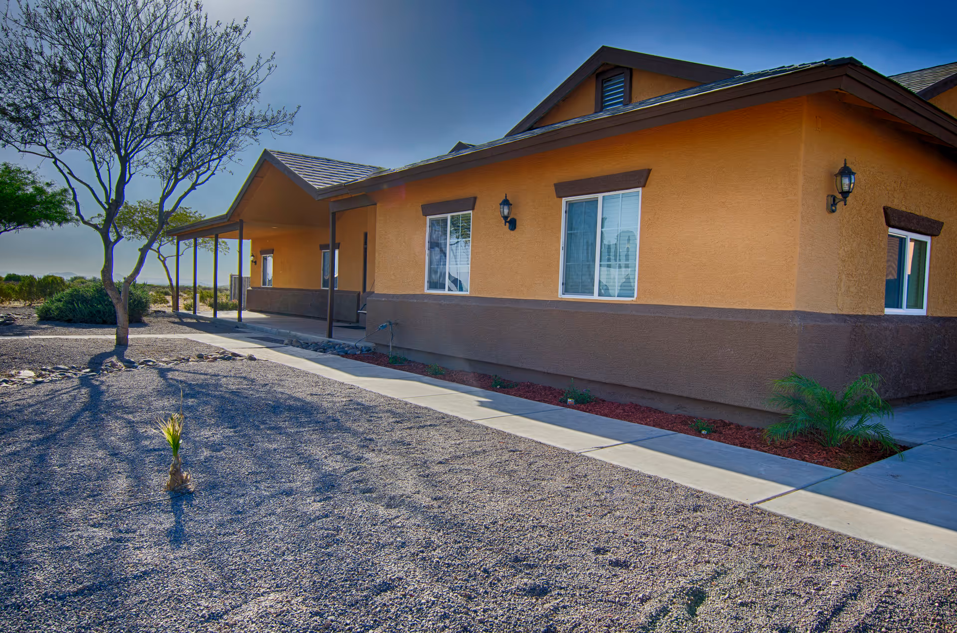 Exterior view of a single-story building with tan and brown walls, several windows, and outdoor wall lanterns. There is a concrete walkway along the side of the building, a small tree, and desert landscaping with gravel and sparse plants under a clear blue sky.