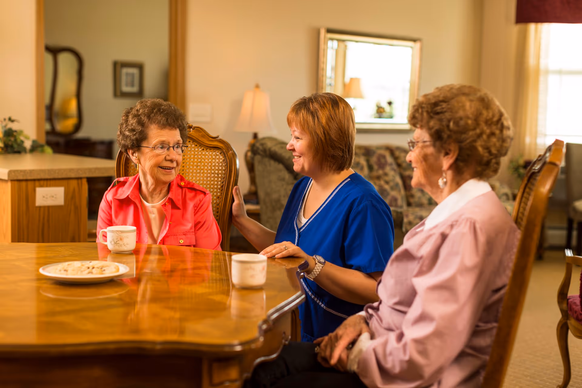 Two elderly women and a caregiver sitting around a wooden table in a warmly lit living room, smiling and engaging in conversation. There are cups and a plate of cookies on the table, with a sofa and lamp in the background.