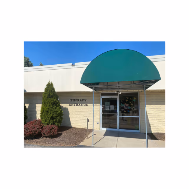 Exterior view of a therapy entrance at a building with a green awning above double glass doors. There are shrubs and a small tree planted in mulch beds next to the entrance. The wall is light-colored with the words 'THERAPY ENTRANCE' displayed beside the door.