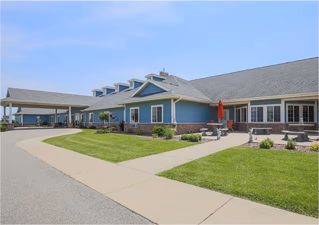 Exterior view of a single-story senior living facility building with blue siding and brick accents. The building has multiple windows, a covered entrance, and a patio area with tables, chairs, and a red umbrella. The surrounding area includes a well-maintained lawn and paved walkways under a clear blue sky.